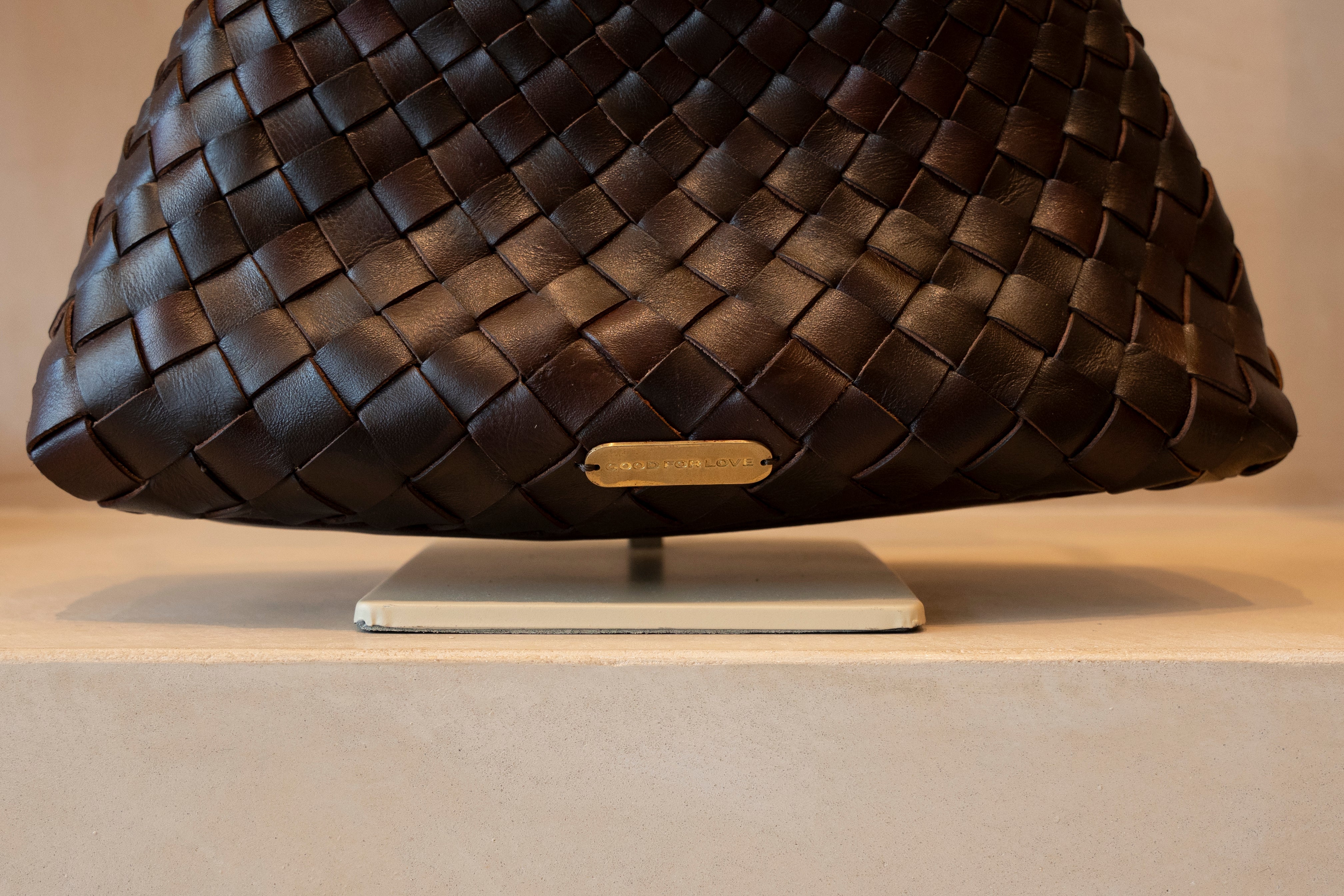 A close-up of a dark brown woven leather handbag displayed on a white stand against a neutral beige background.