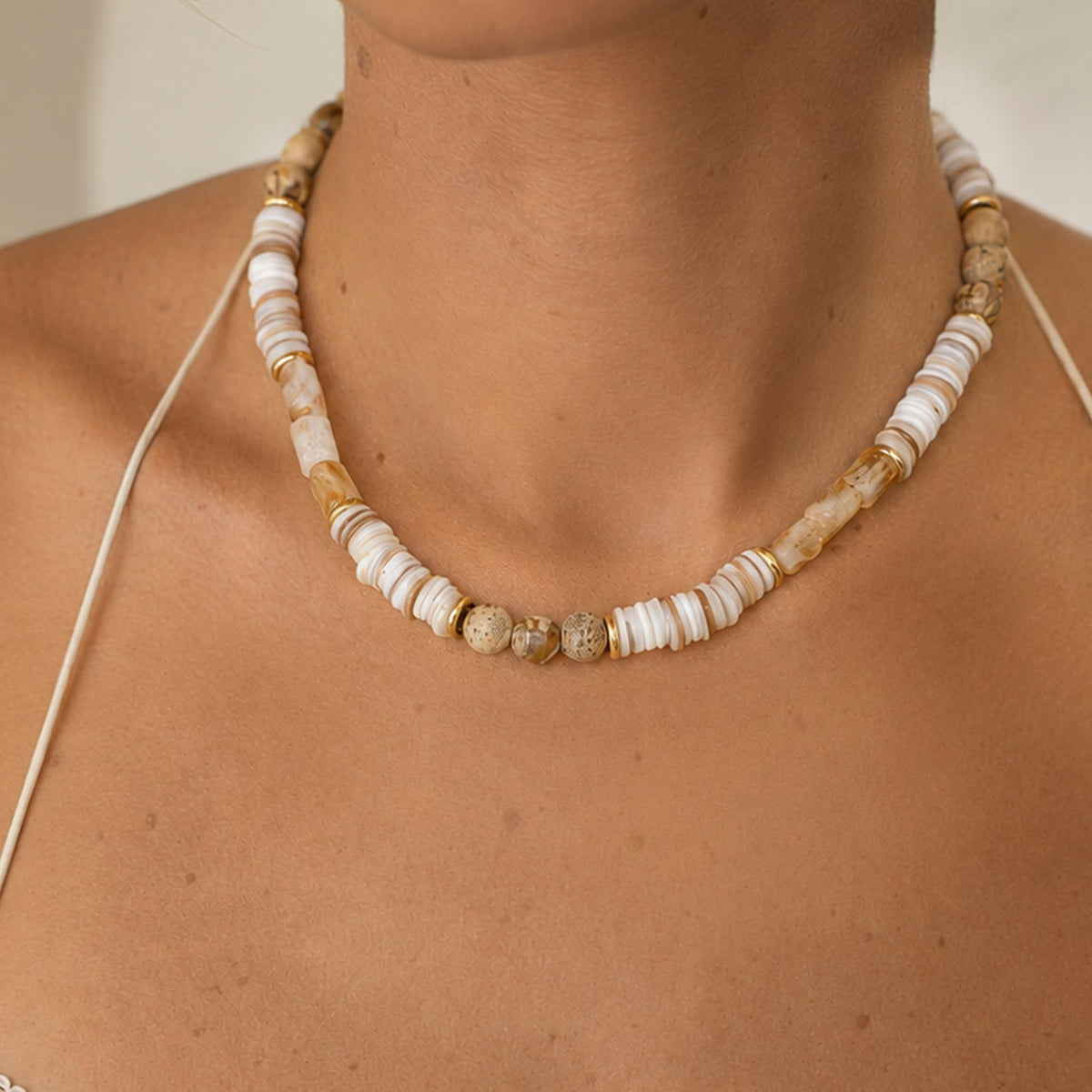 A close-up of a person wearing a beaded necklace made of mostly off-white and beige round beads with a few natural stone accents, paired with a white camisole top.