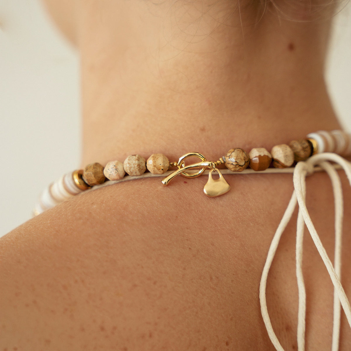 A close-up of a woman's neck and upper back wearing a beaded choker necklace. The necklace features natural wood beads, white beads, and a small gold toggle clasp with a dangling gold heart charm.